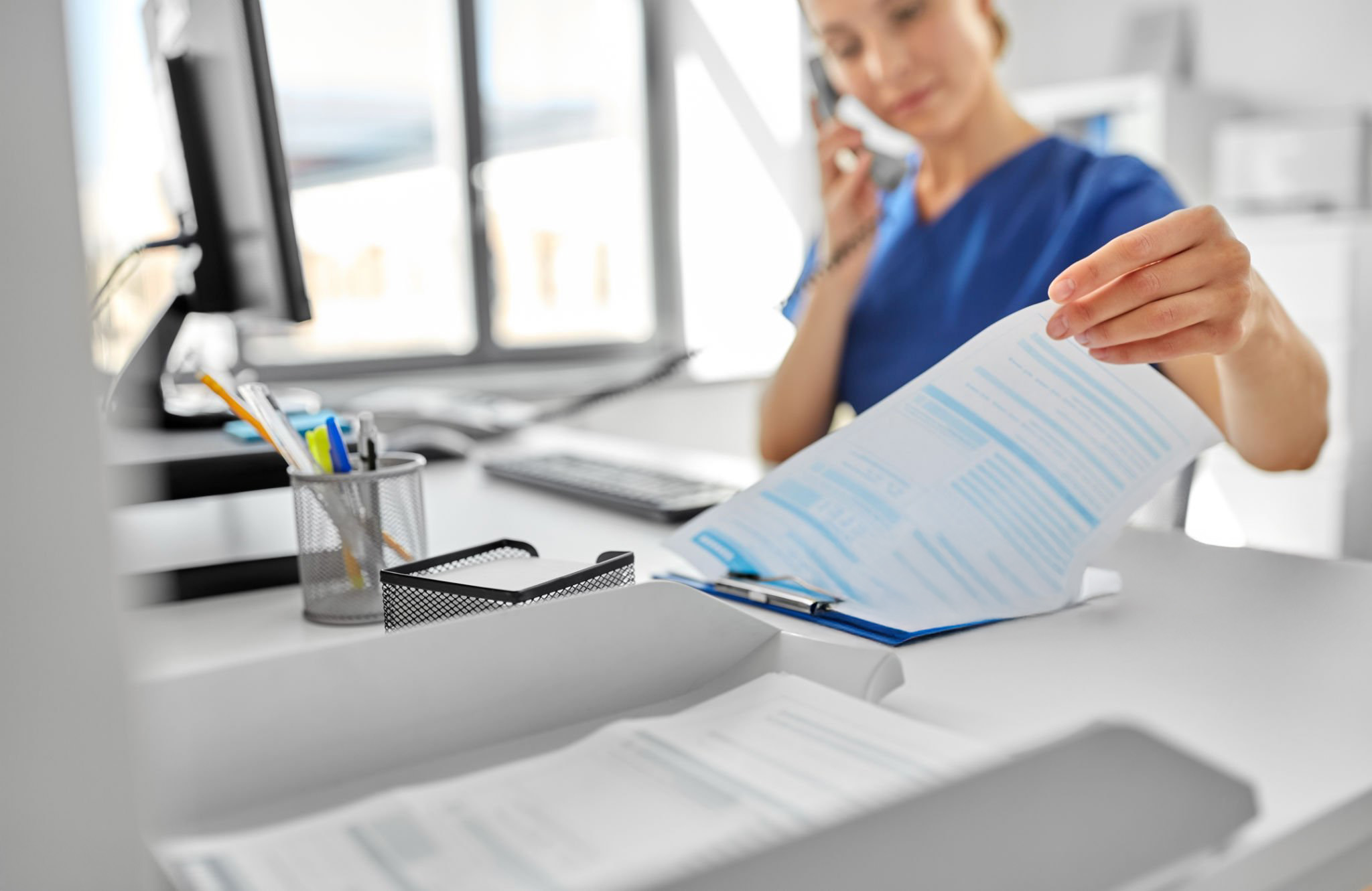 Woman in blue scrubs on the phone sitting at a desk lifting a sheet of paper on a clipboard